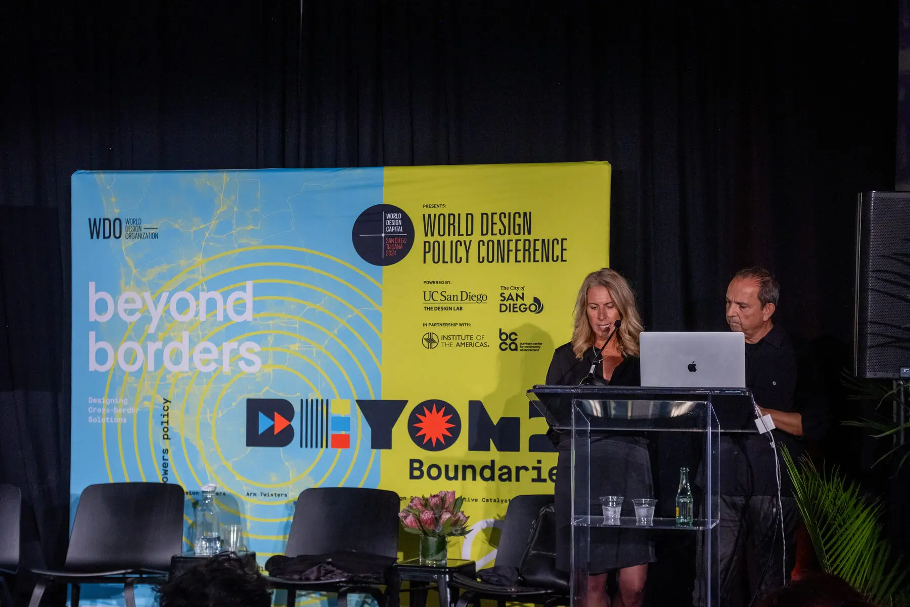 Two speakers at the podium in front of the Beyond Borders backdrop
