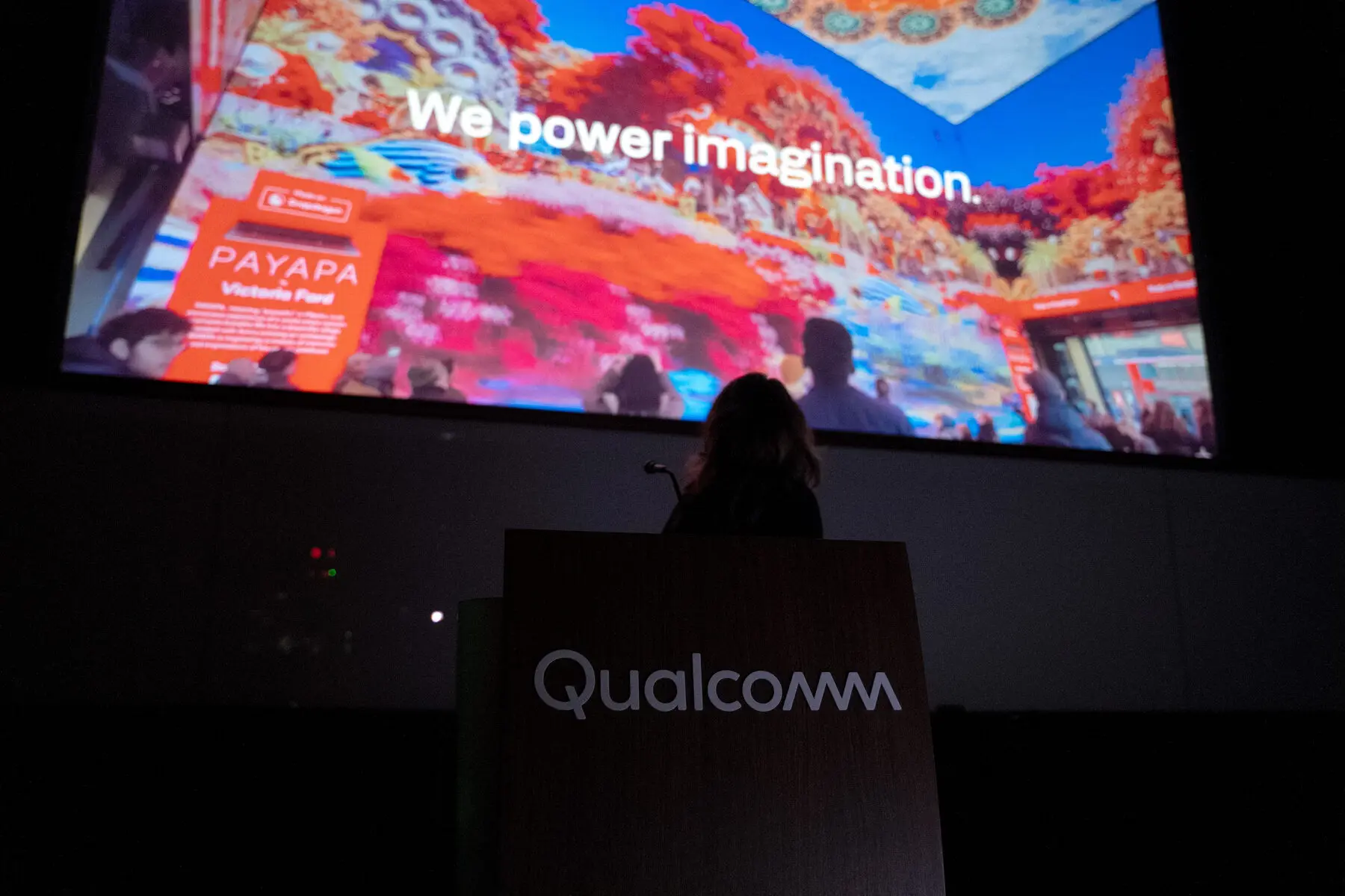 A speaker at a Qualcomm podium in front of a large screen reading We power imagination
